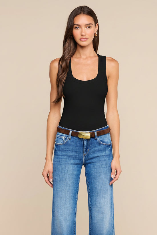A woman modeling a Black ribbed tank top paired with blue jeans and a brown belt, standing against a solid beige studio background.